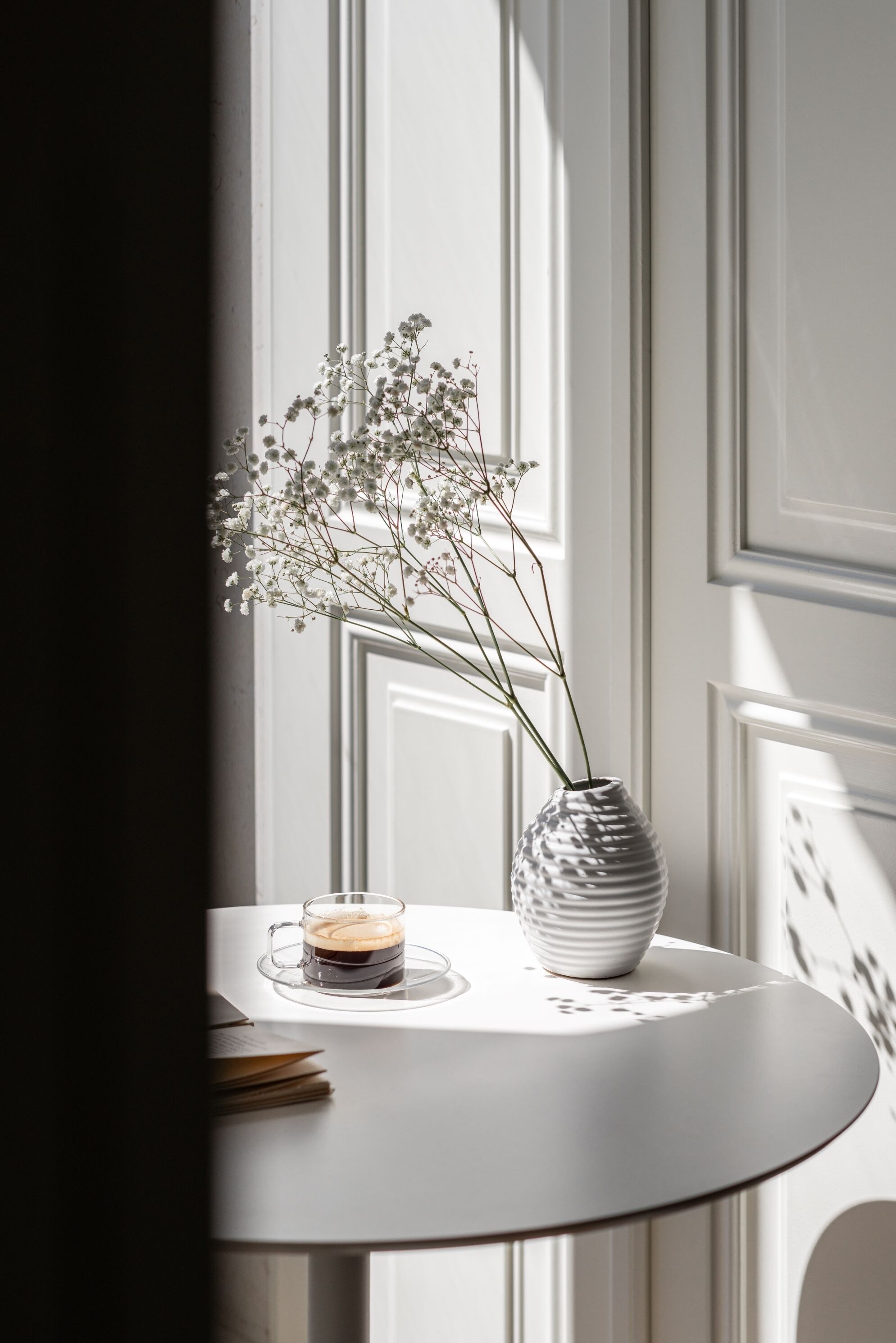 A round white table with a white vase containing white flowers and a glass cup with a beverage.
