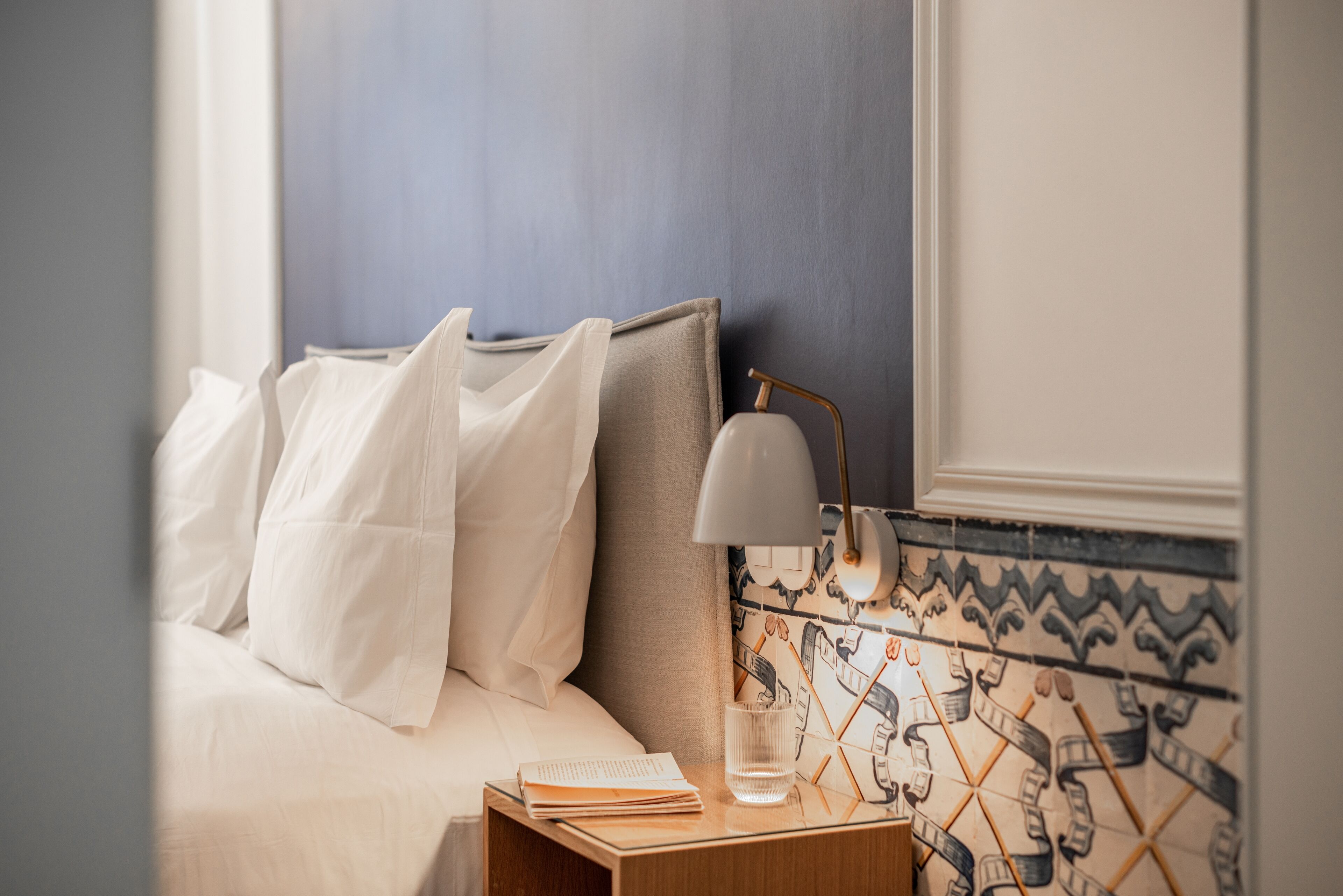 A neatly made bed with white linens, a bedside lamp, a glass of water, and a book on a wooden nightstand.