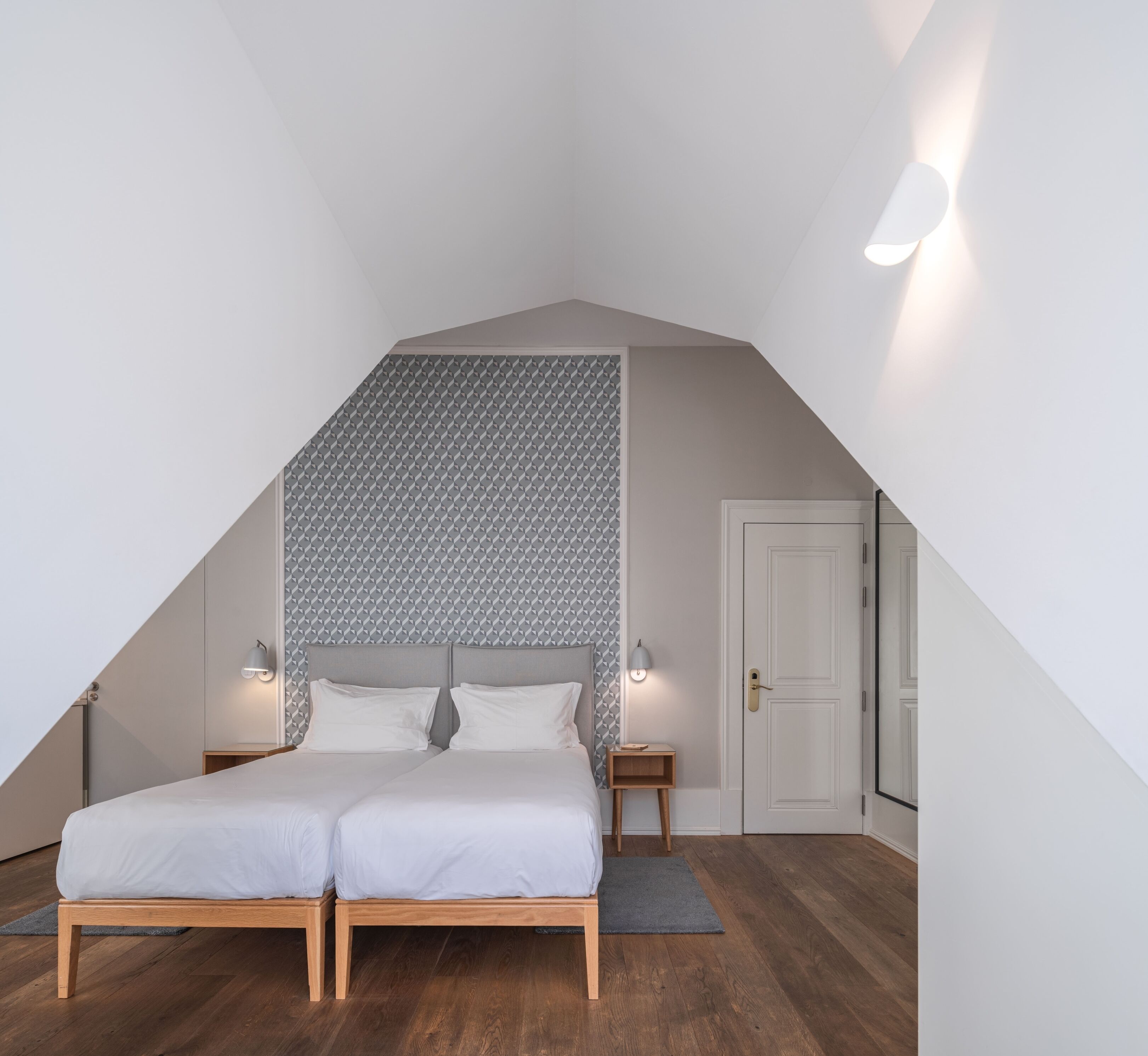 Un dormitorio con techo inclinado, una cama con dos almohadas, lámparas de noche y una puerta.