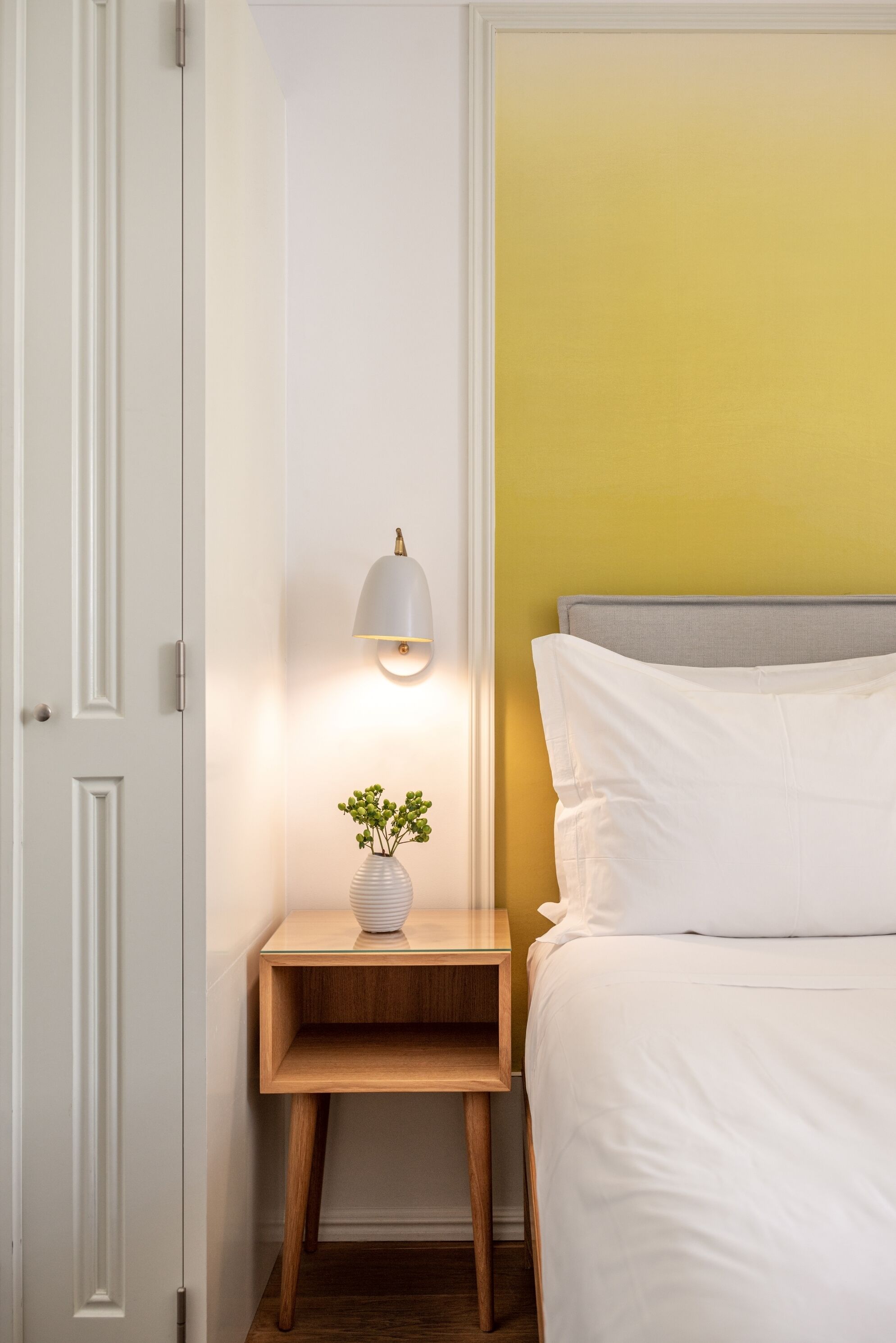Un dormitorio ordenado con cama, mesita de noche, lámpara y una pequeña planta.