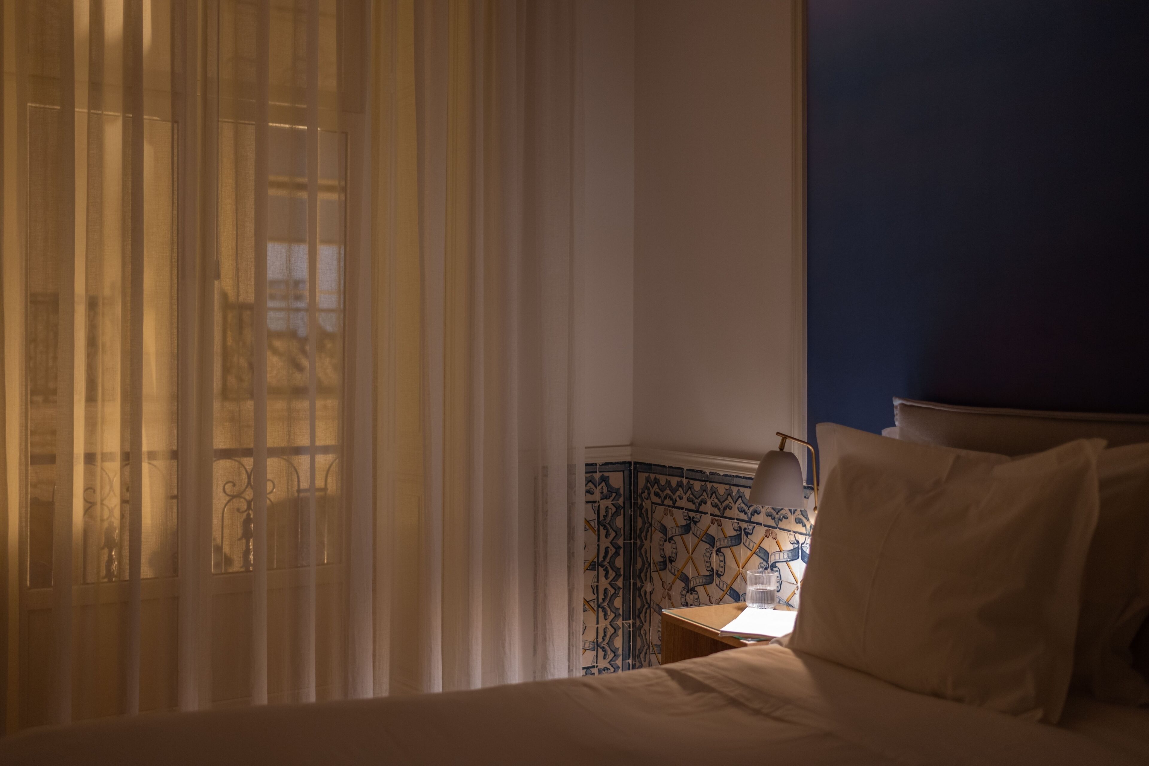A hotel room with a bed, a nightstand, a lamp, and a window with sheer curtains.