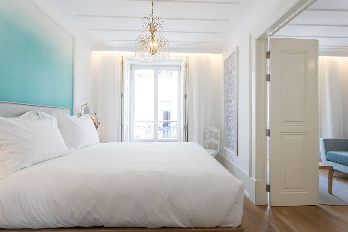 Une chambre à coucher avec un grand lit, un lustre, une vue sur le balcon et une porte donnant sur une autre pièce.