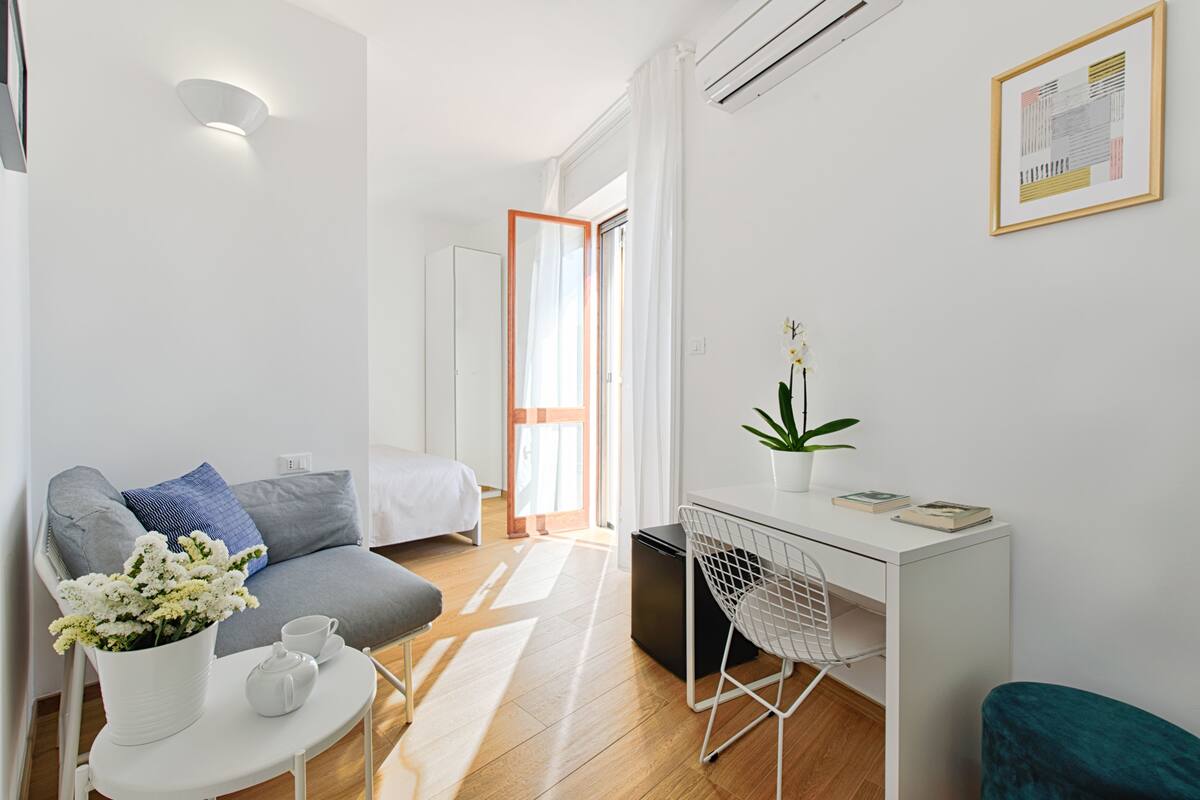 A modern, well-lit room with a bed, a desk, a small table with a vase of flowers, and a potted plant.