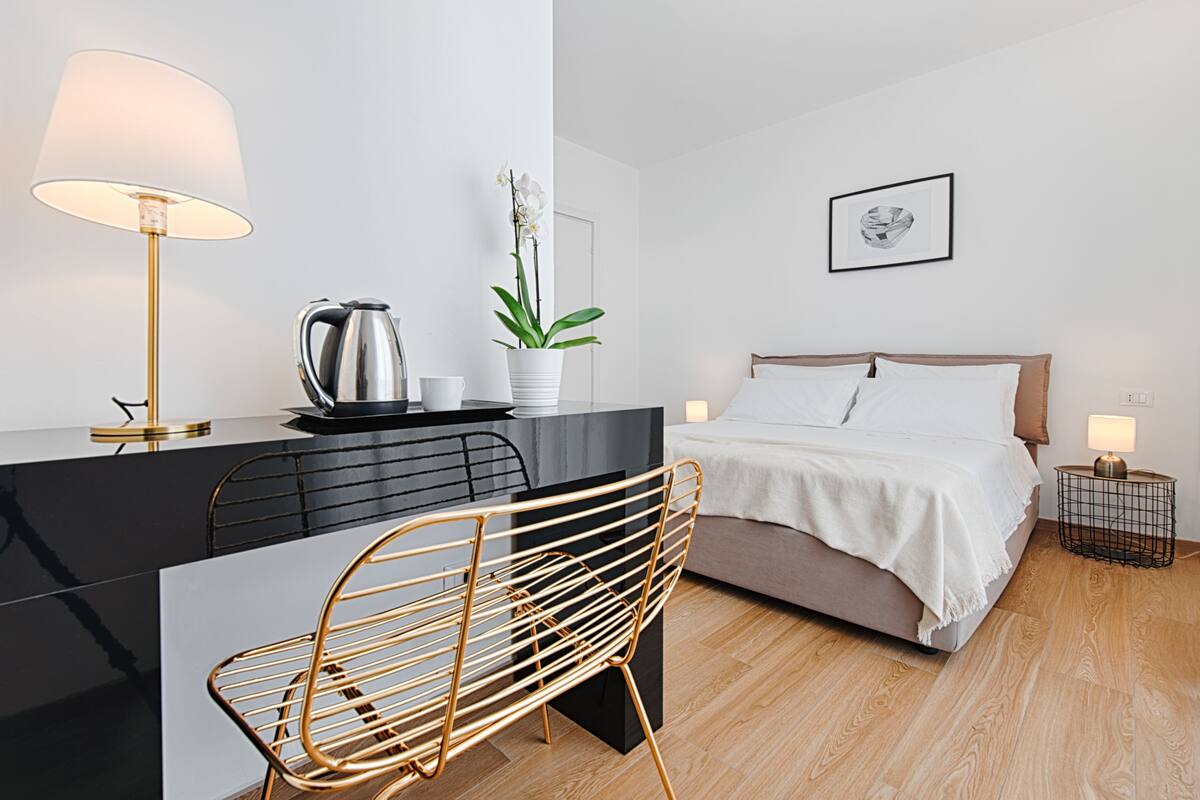 A modern hotel room with a black desk, a gold chair, a bed with white linens, a potted orchid, a framed picture on the wall, and a small side table with a lamp.
