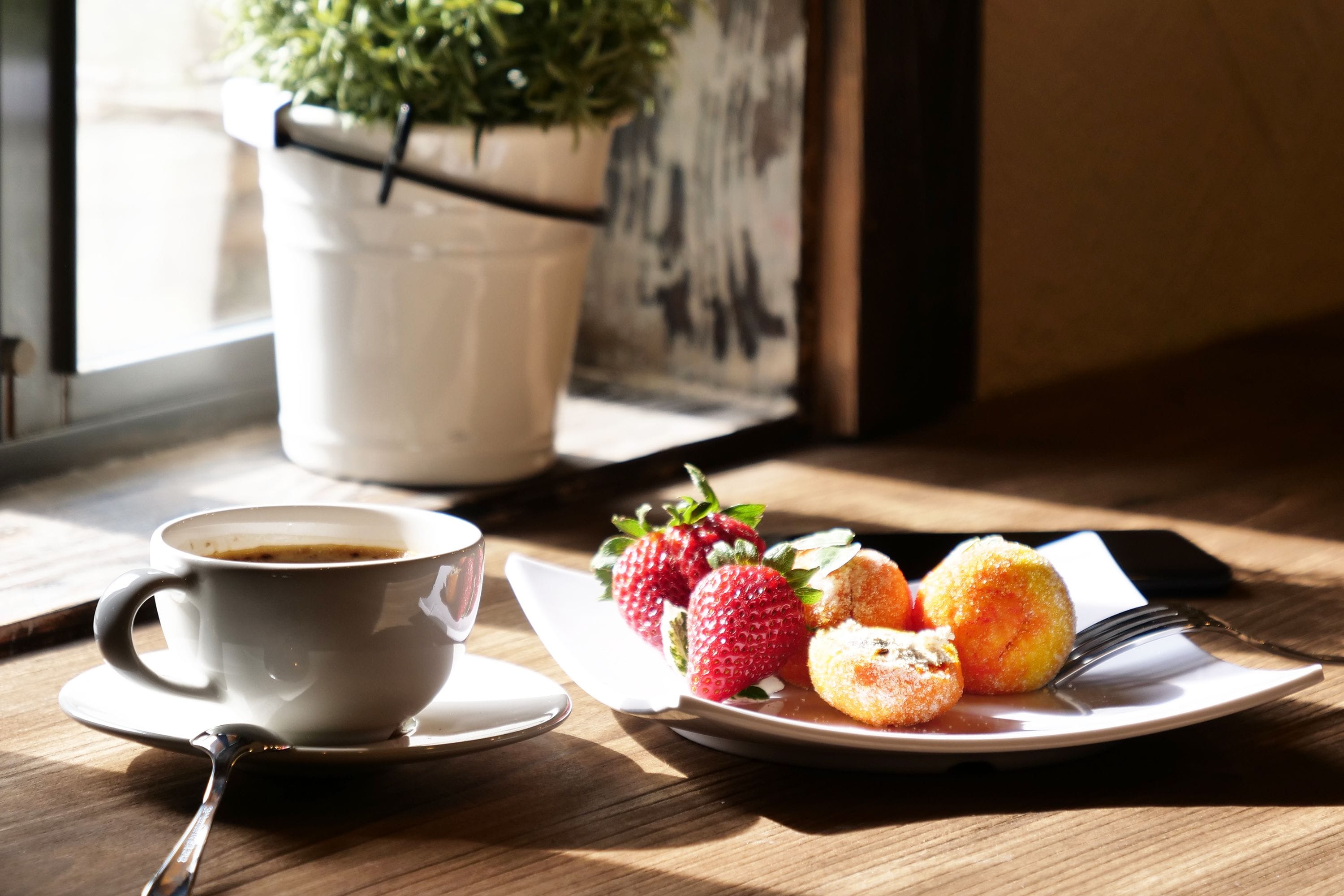 A cup of coffee, strawberries, and pastries on a table near a window with a plant.