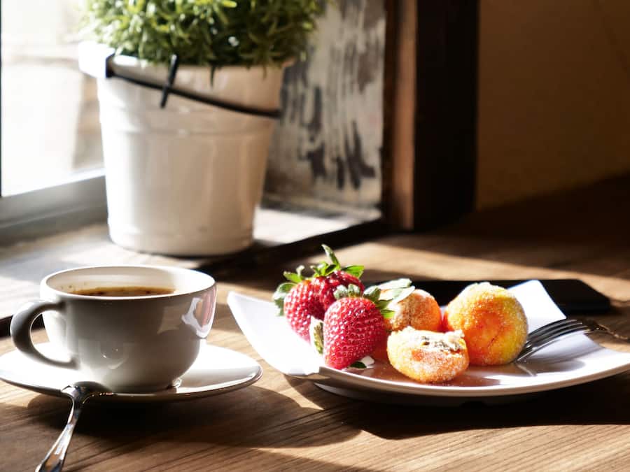 A cup of coffee, strawberries, and pastries on a table near a window with a plant.