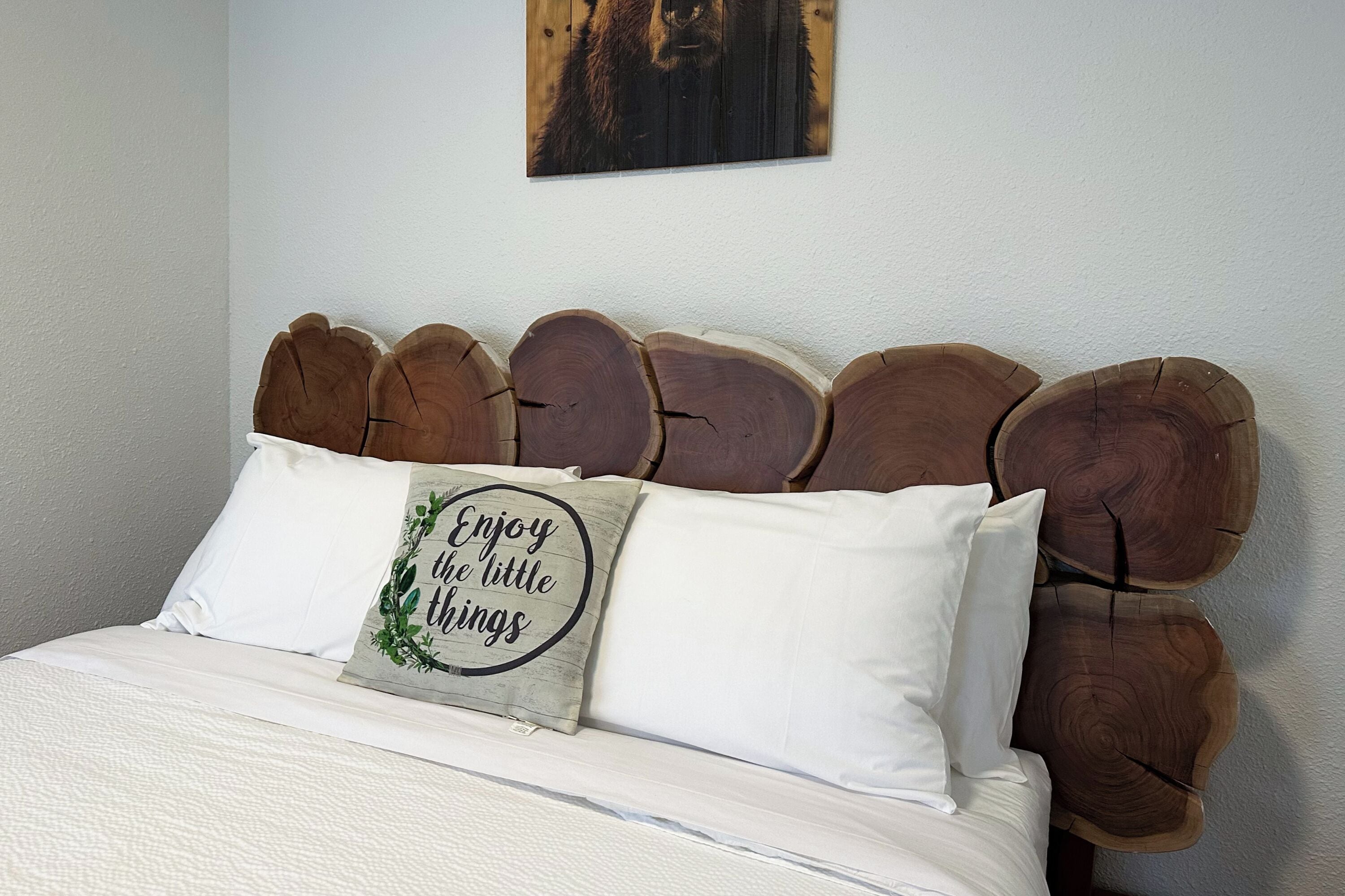 Een bed met een houten hoofdbord, een kussen met een motiverende quote, en een ingelijste foto aan de muur.