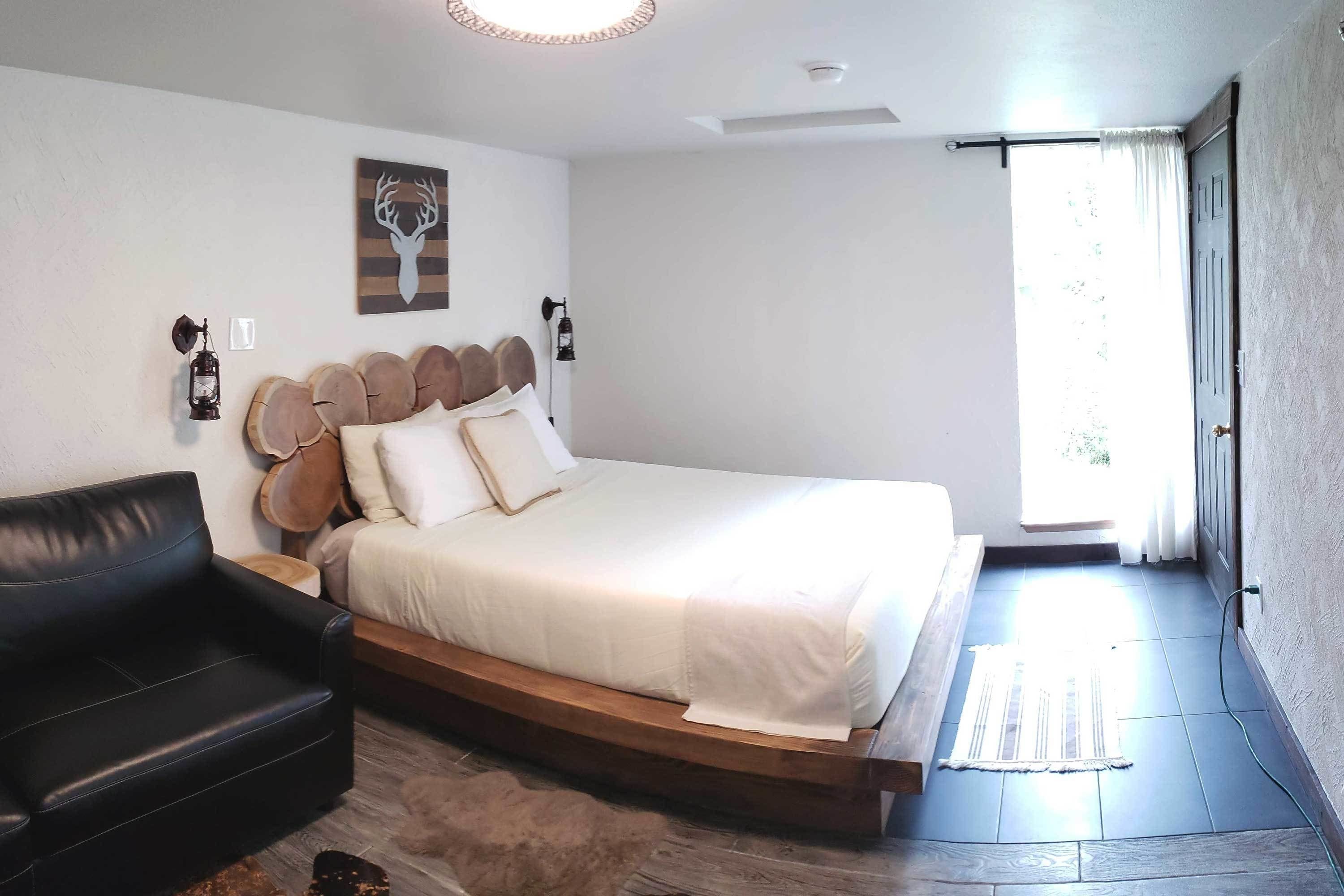 A bedroom with a bed, a black leather sofa, a wooden headboard, a wall-mounted light, and a window with curtains.