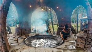 A stone fire pit with a person sitting nearby, surrounded by stone walls and a rustic outdoor setting. - Paradise Village Hotel (Ashford)