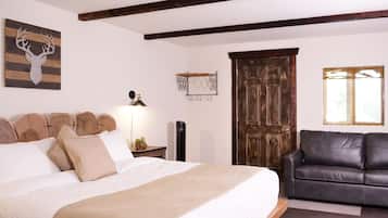 A bedroom with a bed, a wooden headboard, a wall-mounted deer head decoration, a wooden door, a black leather sofa, and a window with blinds.