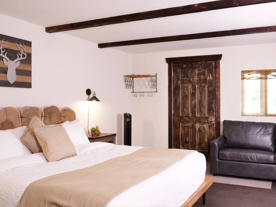 A bedroom with a bed, a wooden headboard, a wall-mounted deer head decoration, a wooden door, a black leather sofa, and a window with blinds.