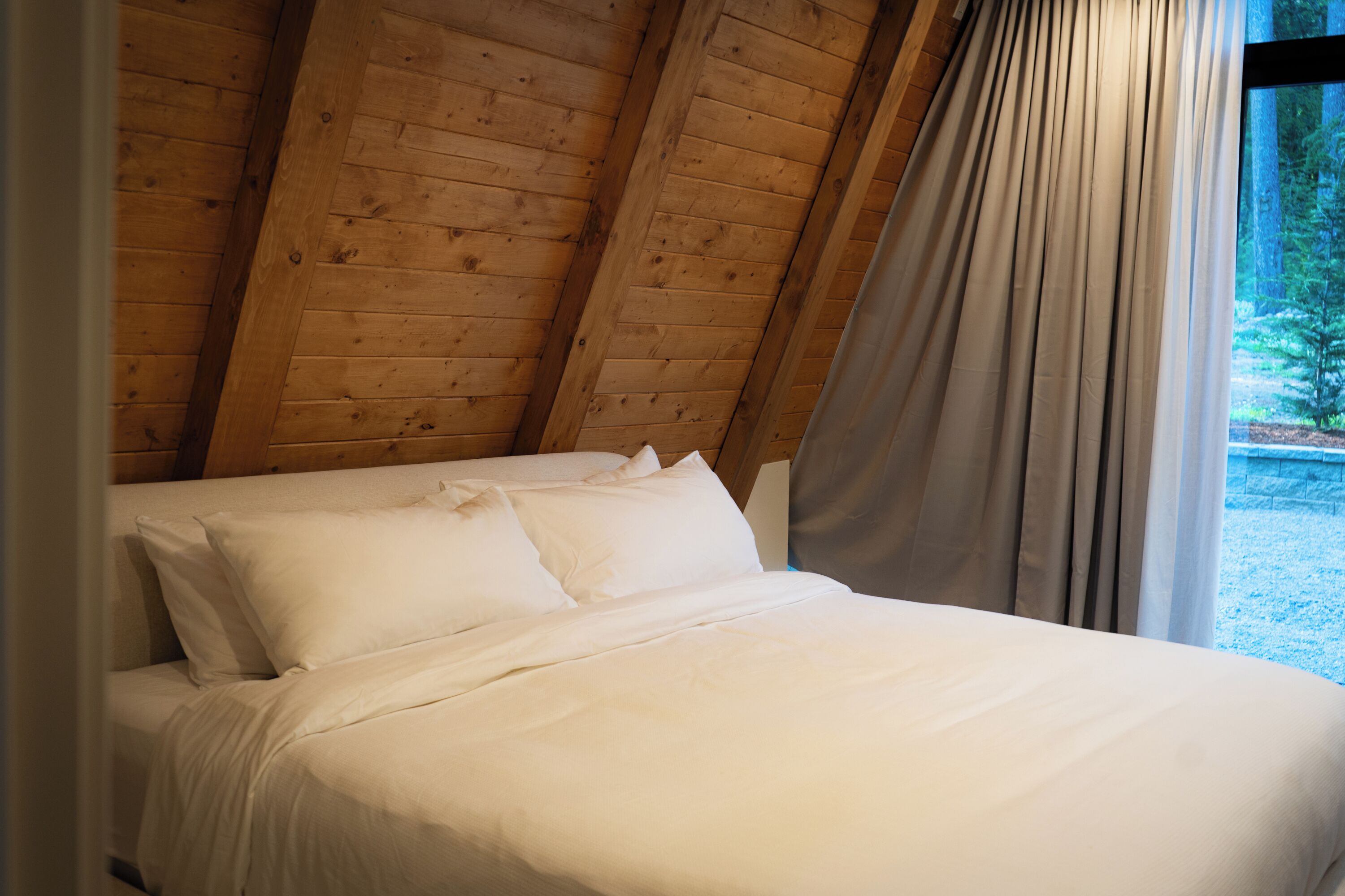 Un lit dans une chambre avec des poutres apparentes en bois au plafond et une vue sur les arbres par la fenêtre.