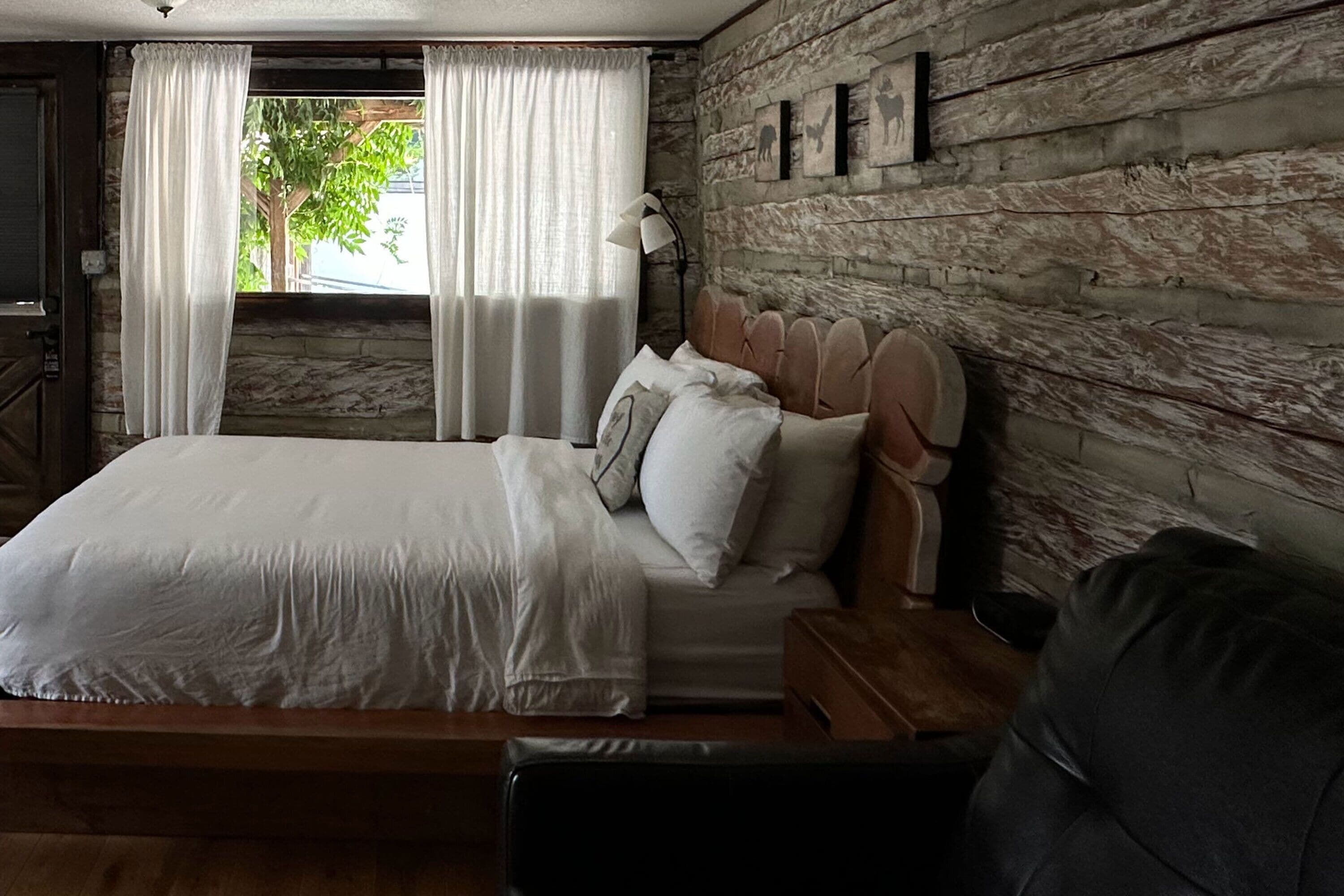 A bedroom with a wooden headboard, a bed with white linens, a nightstand, a lamp, and a view of the outdoors through a window with curtains.