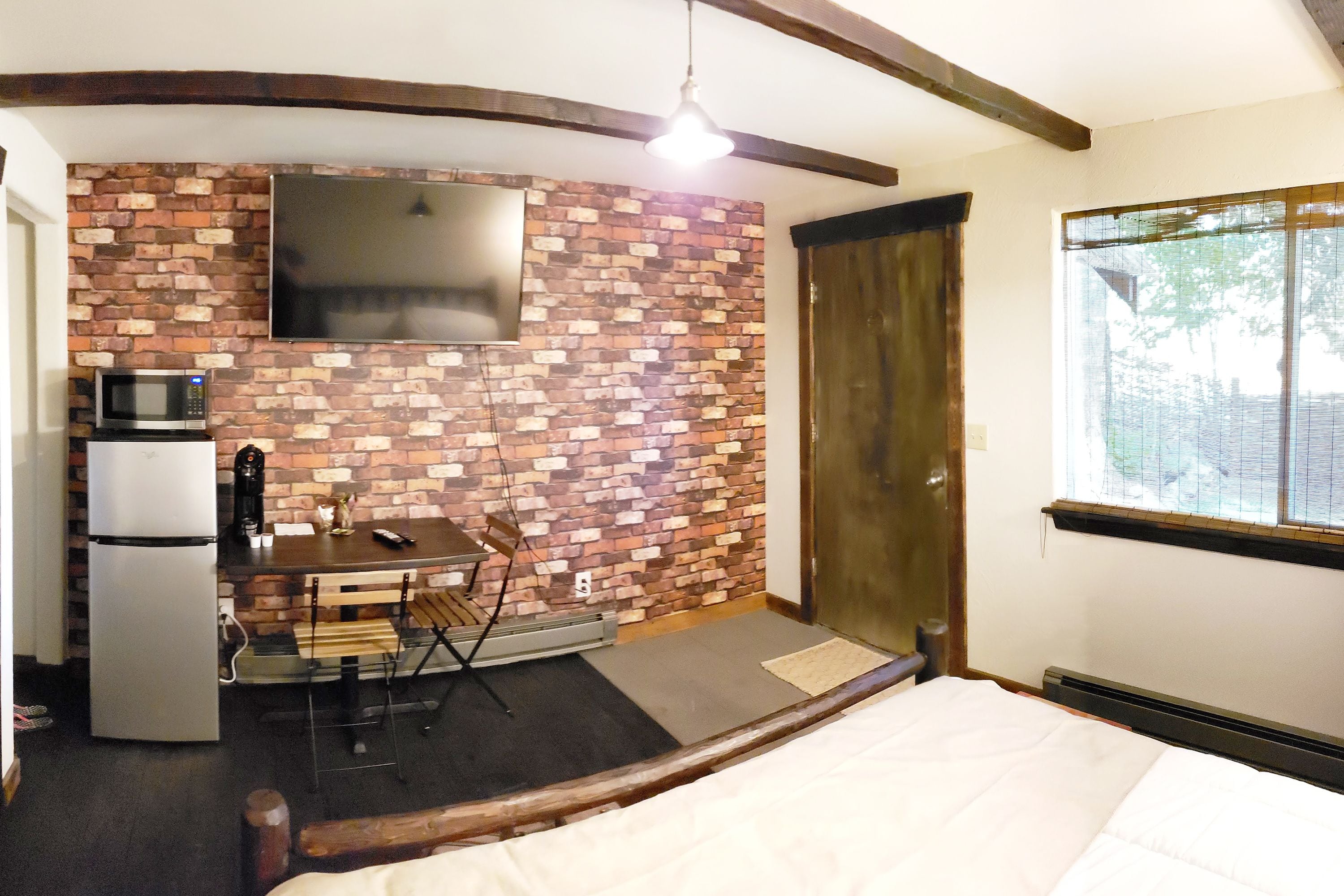 A compact room with a brick wall, a flat-screen TV, a small kitchenette with a fridge, microwave, and coffee maker, a wooden bench, and a bed with a white comforter.