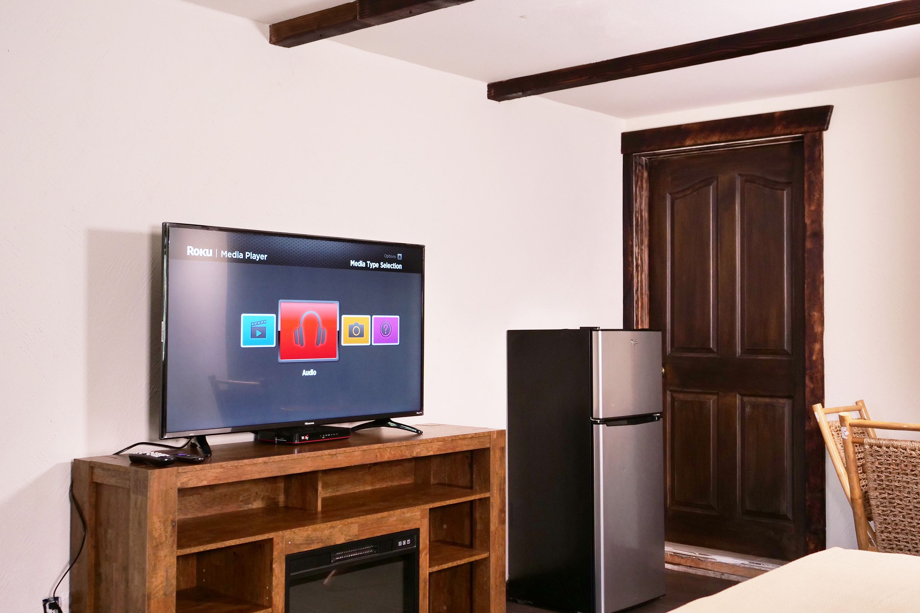 A television on a wooden stand with a digital interface, a black refrigerator, and a wooden door in a room with white walls.
