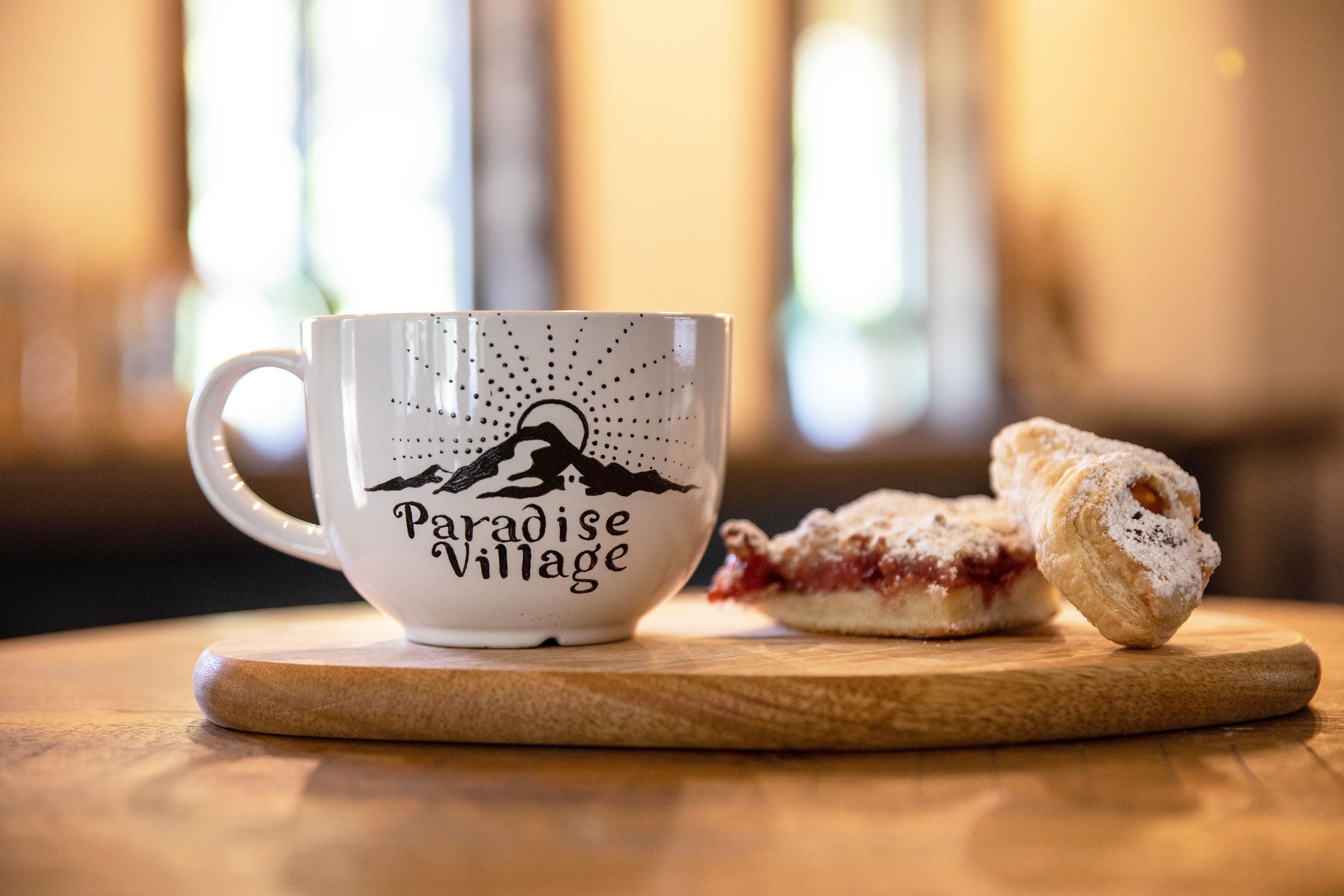 A white mug with "Paradise Village" text and a mountain design, placed on a wooden coaster with a pastry beside it.