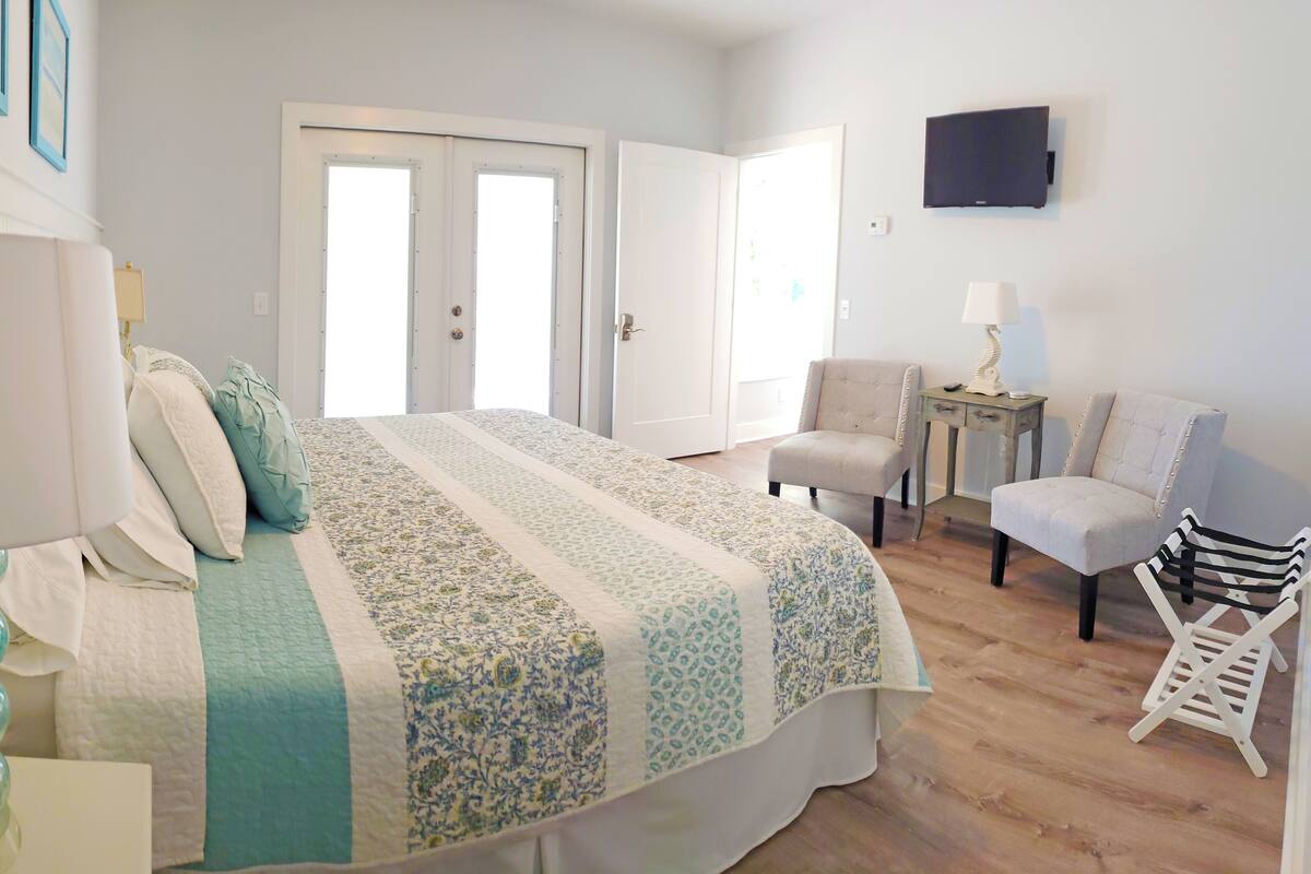 A bedroom with a bed, two doors, a chair, a small table, and a TV on the wall.