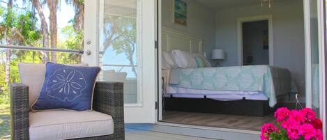 Une chaise en osier avec un coussin et un oreiller décoratif, un lit recouvert d’une couvre-lit verte et blanche, et une plante en pot aux fleurs roses, le tout sur une terrasse en bois.