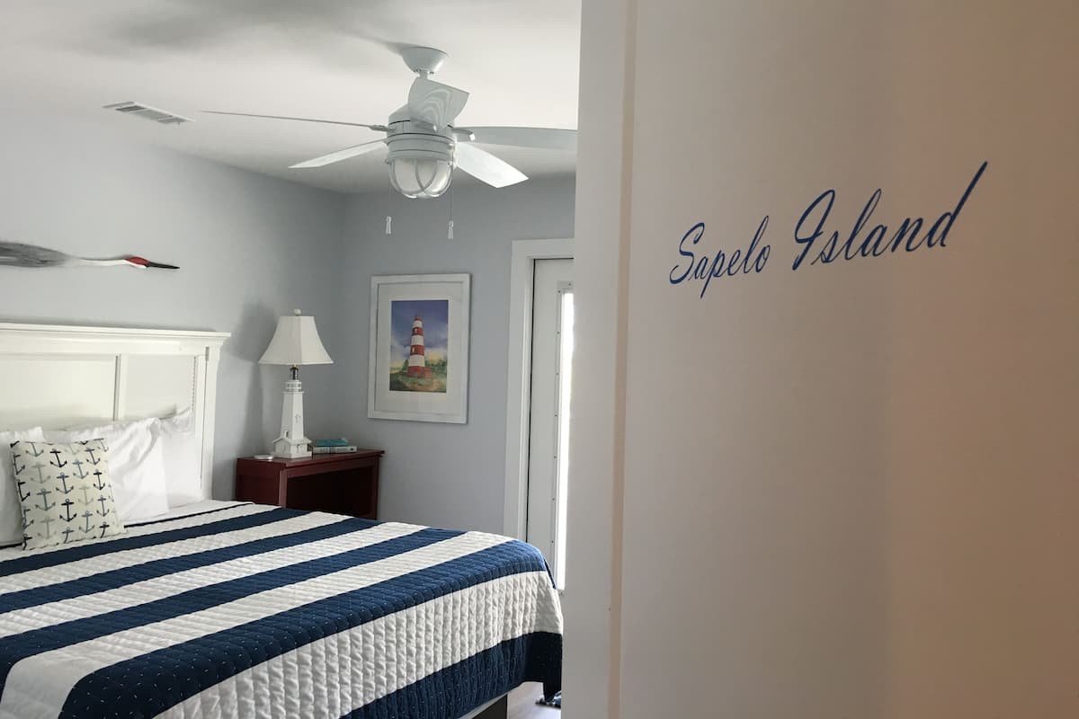 A bedroom with a bed, a ceiling fan, a nightstand, a lamp, a framed picture, and a wall with the text "Sapelo Island".