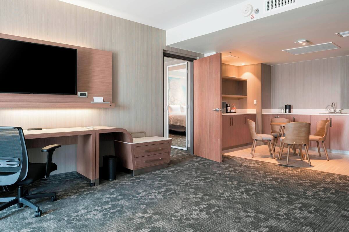 A modern hotel room with a desk, chair, and a TV. There is a dining area with chairs and a table.