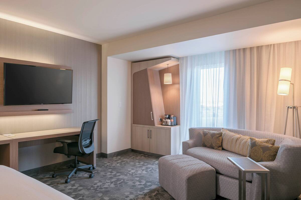 A hotel room with a flat-screen TV, a desk, a chair, a sofa, and a lamp.