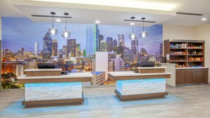 Zona de recepción de hotel con dos mostradores, un mural con vista urbana, iluminación colgante y un estante para aperitivos.