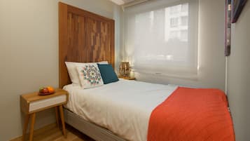 A bedroom with a wooden headboard, a bed with white bedding and a red throw, a bedside table with a fruit bowl, and a window with sheer curtains.