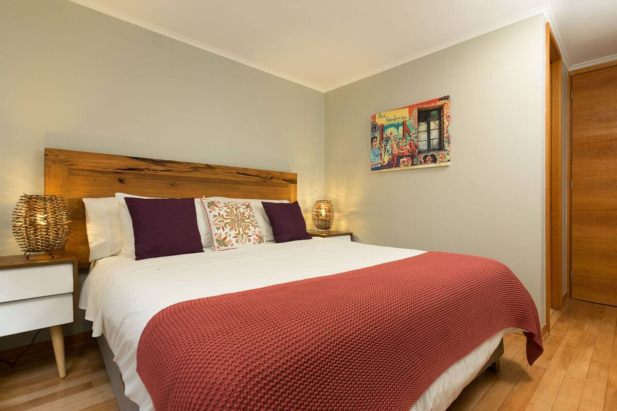 Ein ordentlich bezogenes Bett mit roter Decke, zwei weißen Kissen und einem Holzkopfende. Darüber an der Wand hängt ein farbenfrohes Bild, und zur rechten Seite befindet sich eine Holztür.