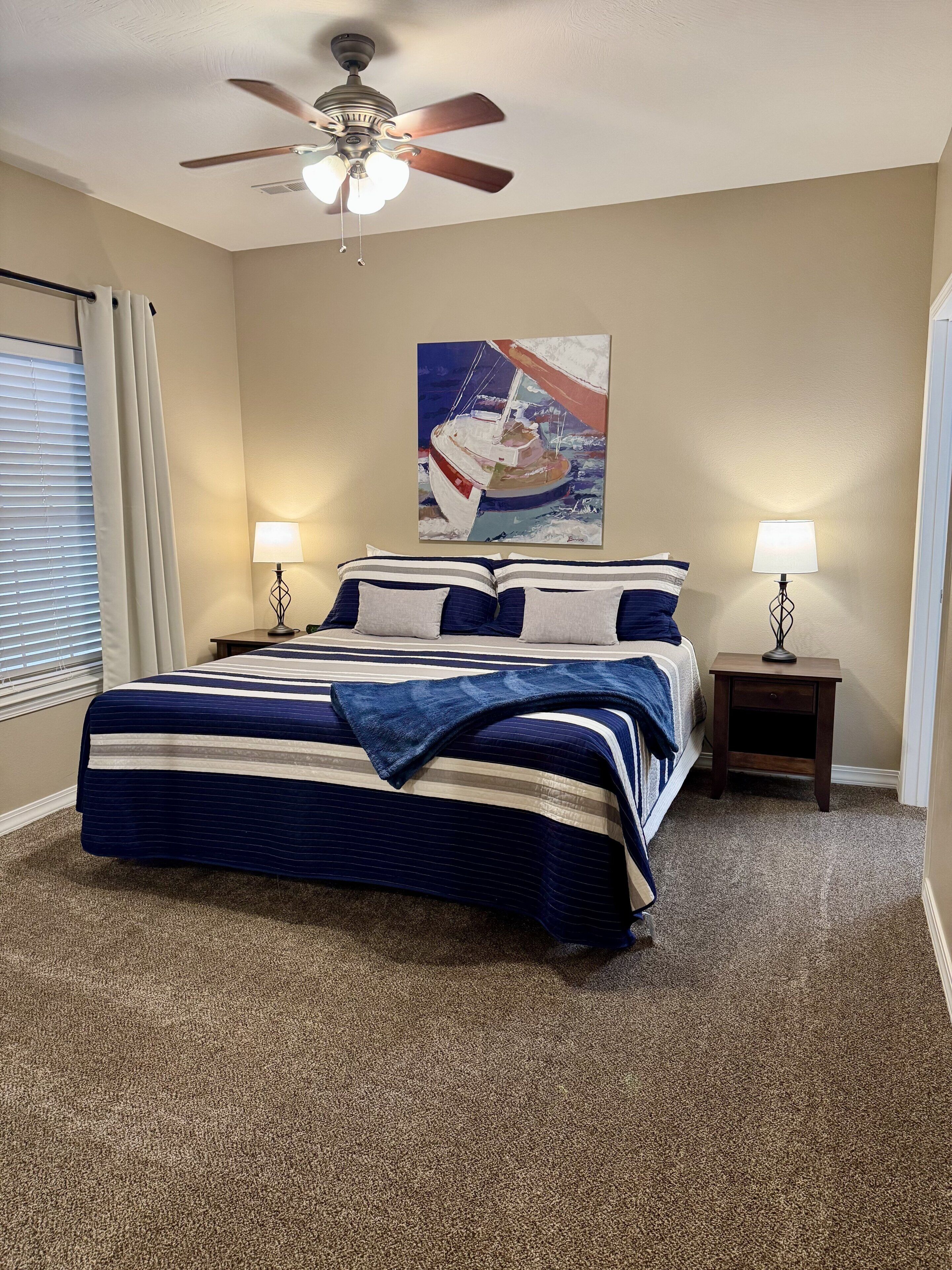 A bedroom with a bed, two bedside tables, a ceiling fan, a painting, and a window with curtains.