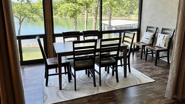 Un comedor con una mesa y sillas, un ventilador de techo, ventanas amplias con vistas a árboles y agua, y piso de madera.