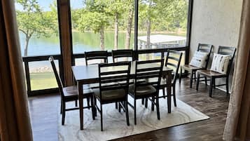 A dining area with a table and chairs, a ceiling fan, large windows with a view of trees and water, and a wooden floor.