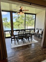 Un comedor con una mesa y sillas, un ventilador de techo, ventanas amplias con vistas a árboles y agua, y piso de madera.