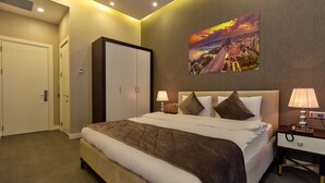 A hotel room with a large bed, a wardrobe, a desk, and a cityscape painting on the wall. - Aksent Hotel (Baku)