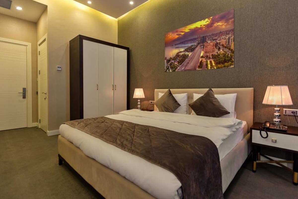 A hotel room with a large bed, two bedside tables with lamps, a wardrobe, and a cityscape painting on the wall.