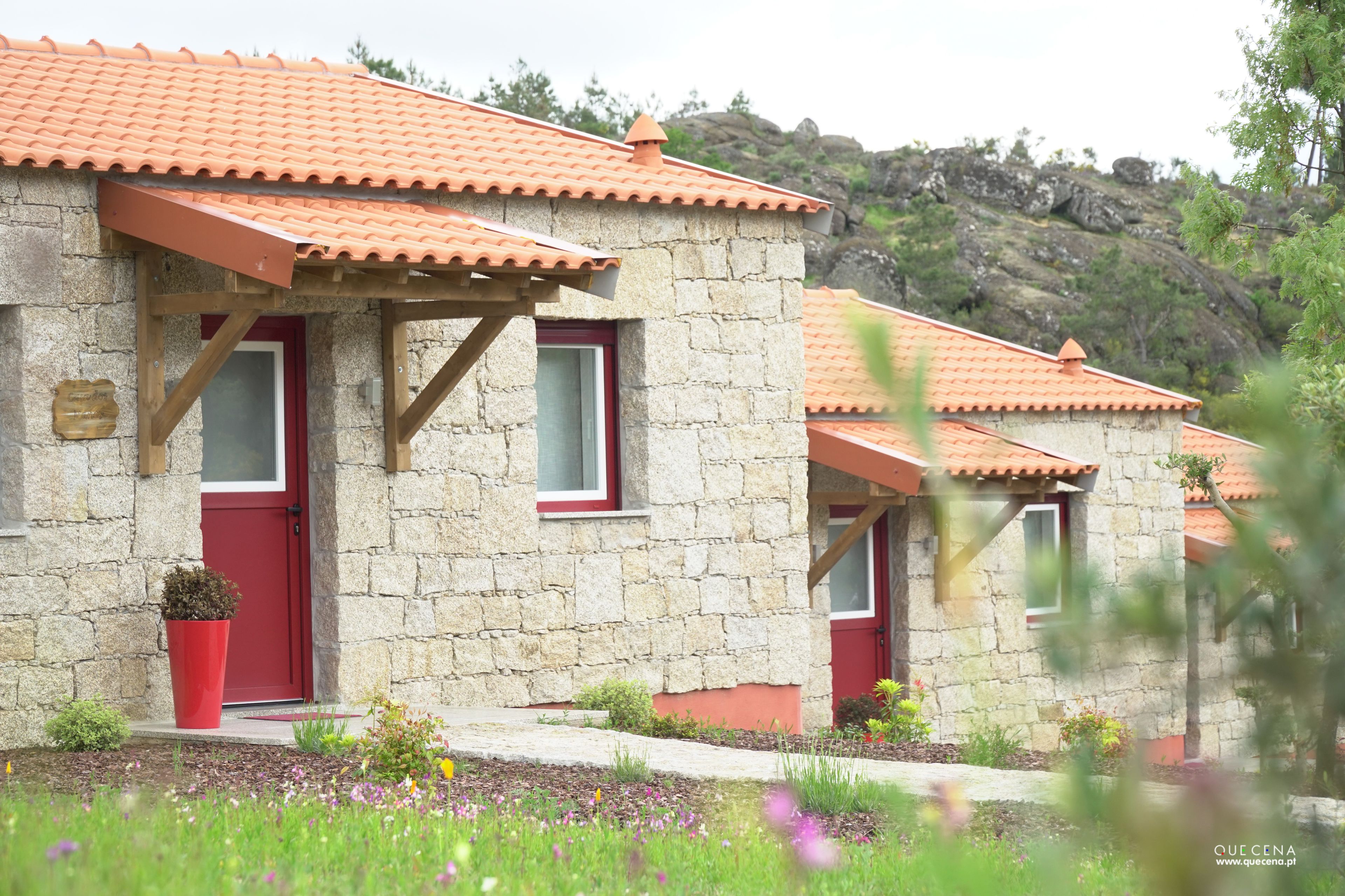 A stone building with a red door and windows, surrounded by a garden with flowers and a hilly landscape in the background.