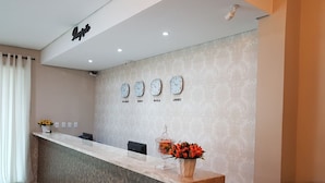 A hotel reception area with a marble counter, clocks displaying different time zones, and a vase of red flowers. - Hotel Loureiro (Rio Branco)