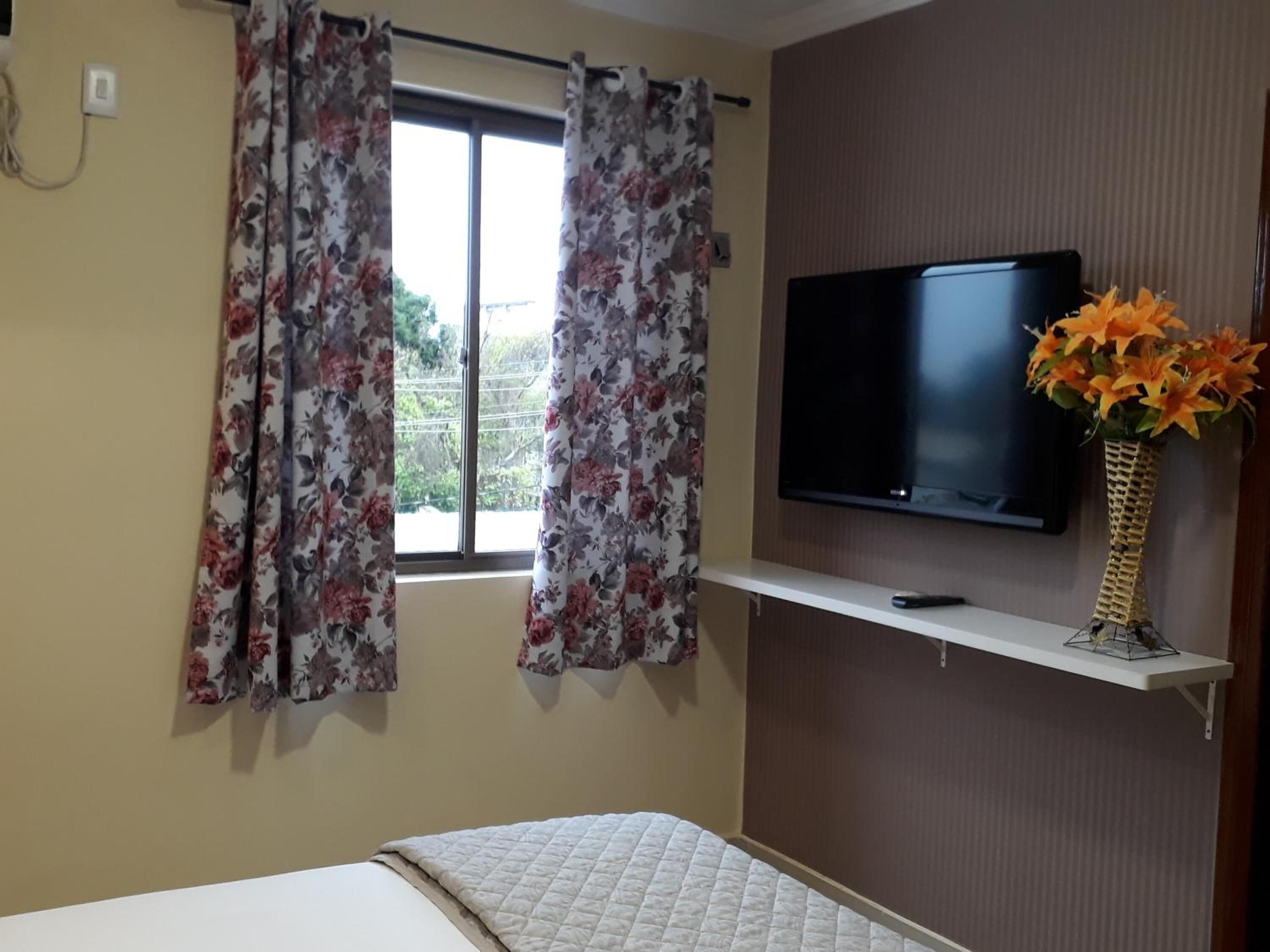 A bedroom with a bed, a TV mounted on the wall, and a window with floral curtains.