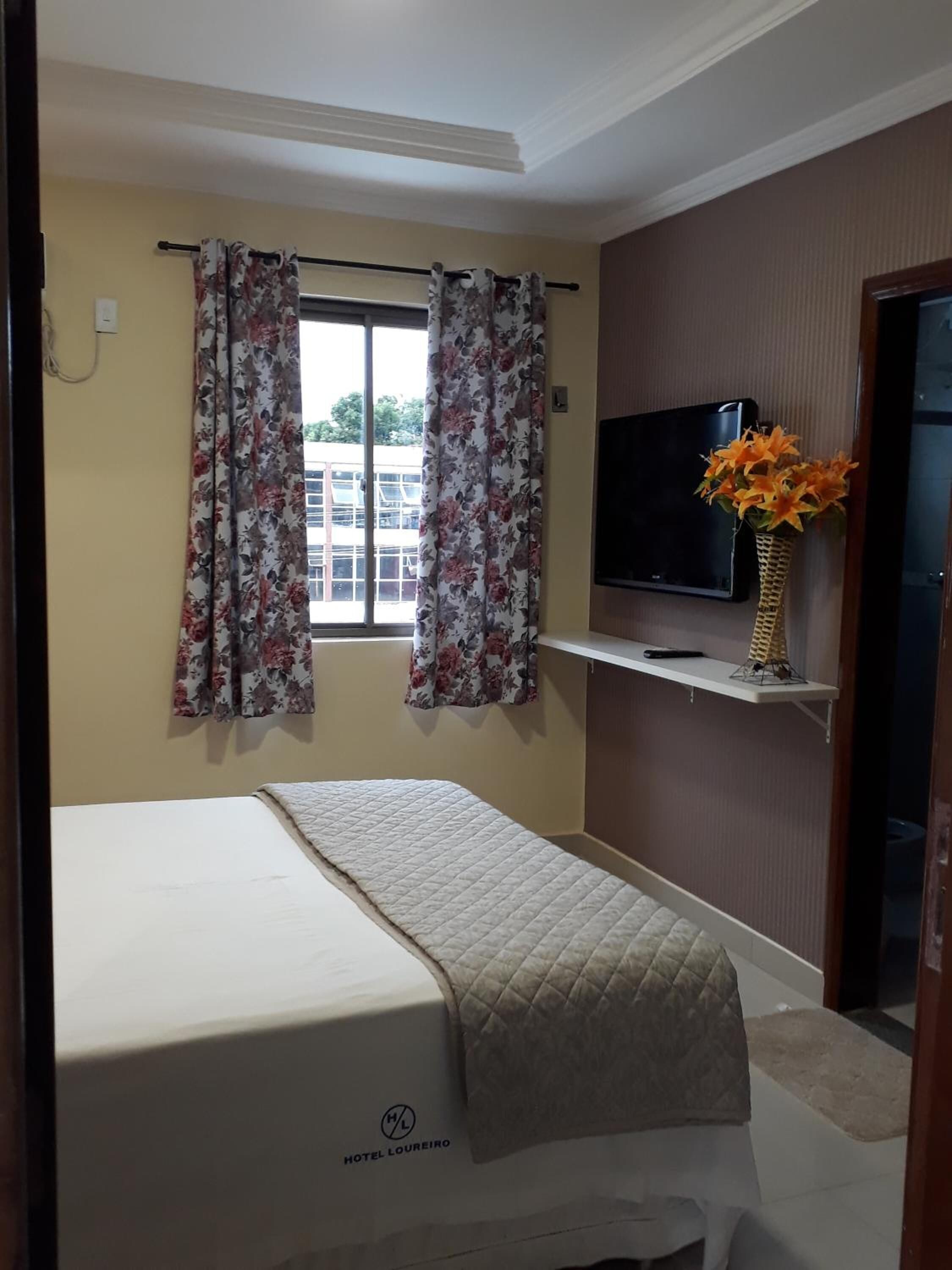 A hotel room with a bed, a television on a shelf, and a window with floral curtains.