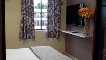 A hotel room with a bed, a television on a shelf, and a window with floral curtains.