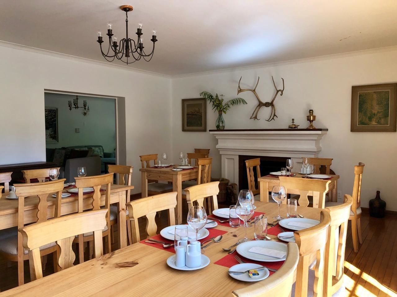A dining area with wooden tables and chairs, a chandelier, and a fireplace with antlers on the wall.