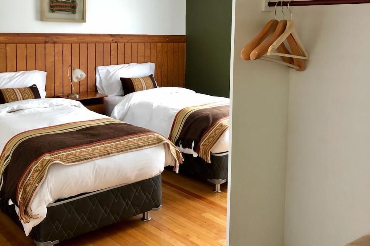 A hotel room with two beds, wooden flooring, a hanging light fixture, and a coat rack with hangers.