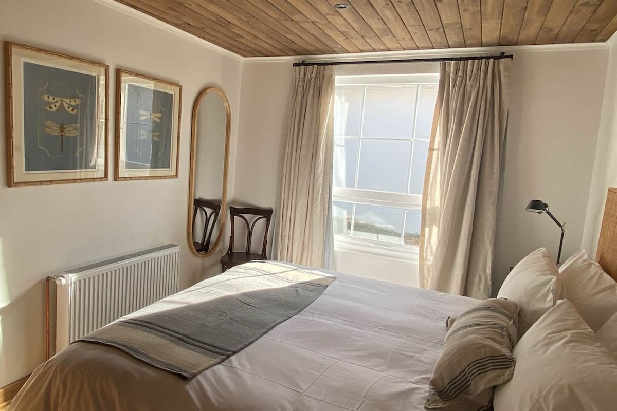 Une chambre à coucher avec un lit, un radiateur, une fenêtre avec des rideaux, un miroir et deux tableaux encadrés accrochés au mur.