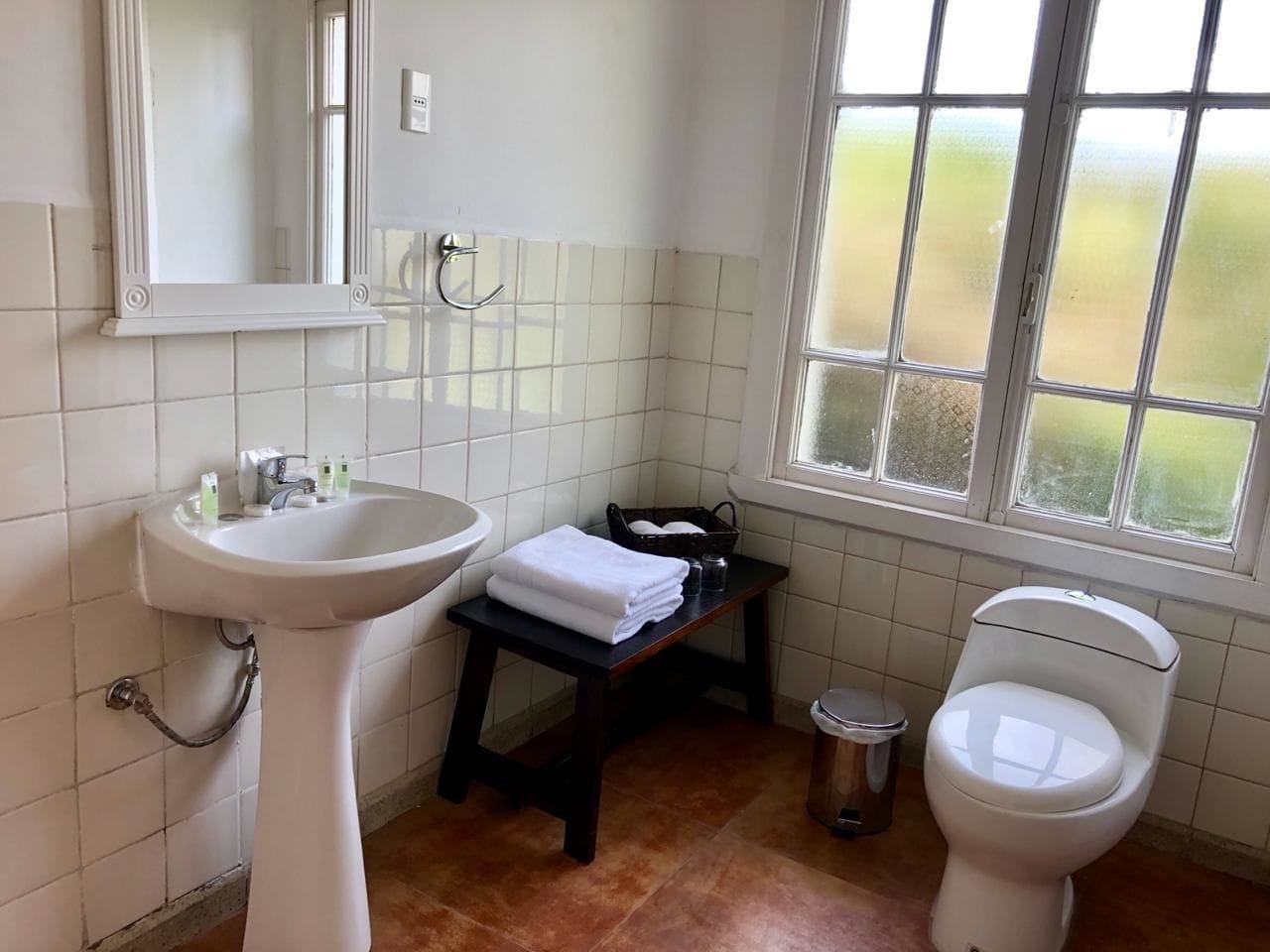 Un baño con lavamanos pedestal, inodoro y una mesita con una toalla.
