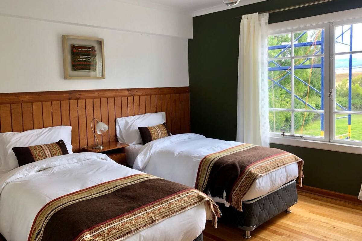 Une chambre avec deux lits, une tête de lit en bois, un tableau encadré au mur et une fenêtre donnant sur un espace verdoyant et un terrain de jeux.