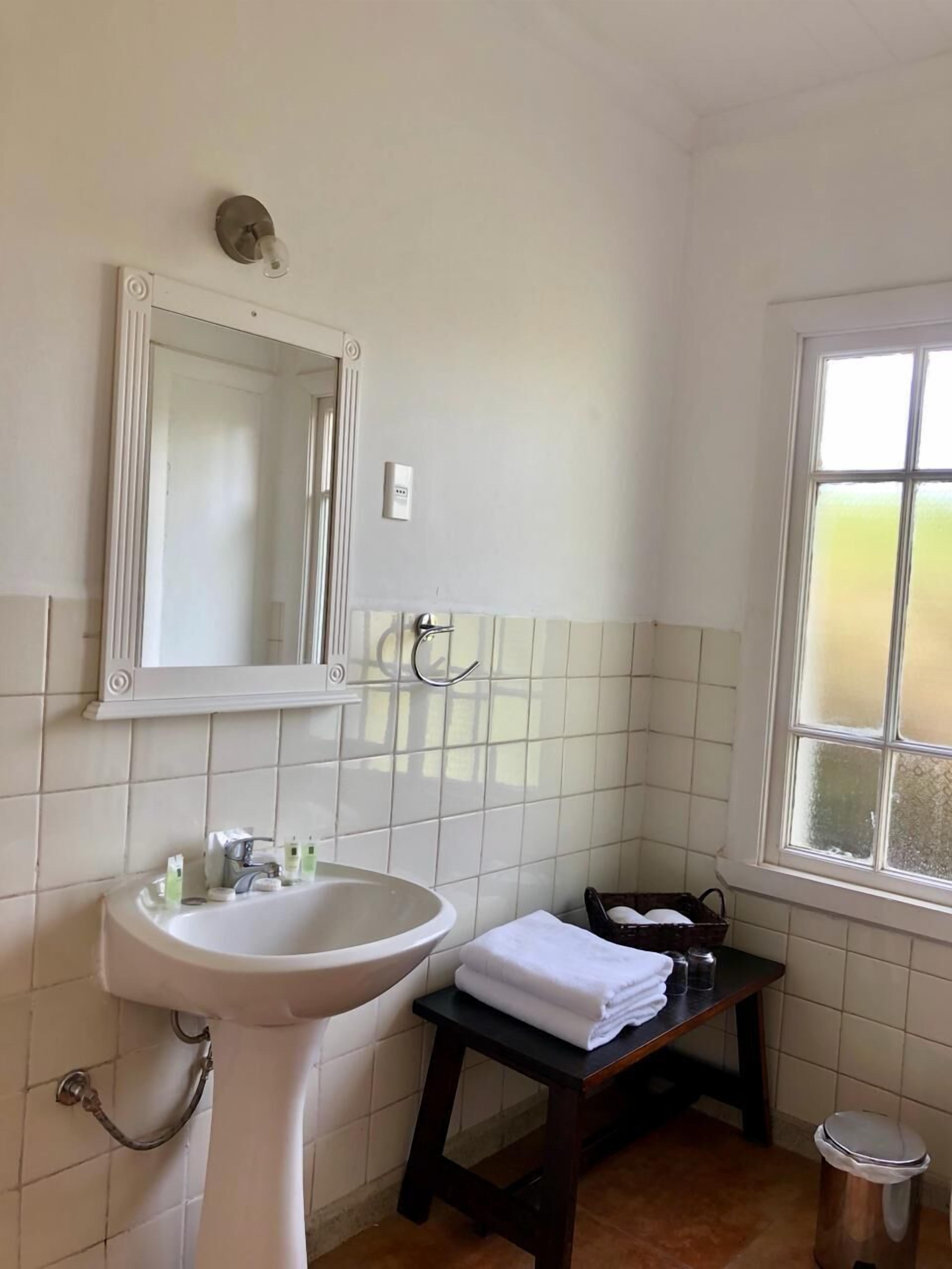 Un baño con lavamanos pedestal, espejo, toallero, ventana y un pequeño cubo de basura.