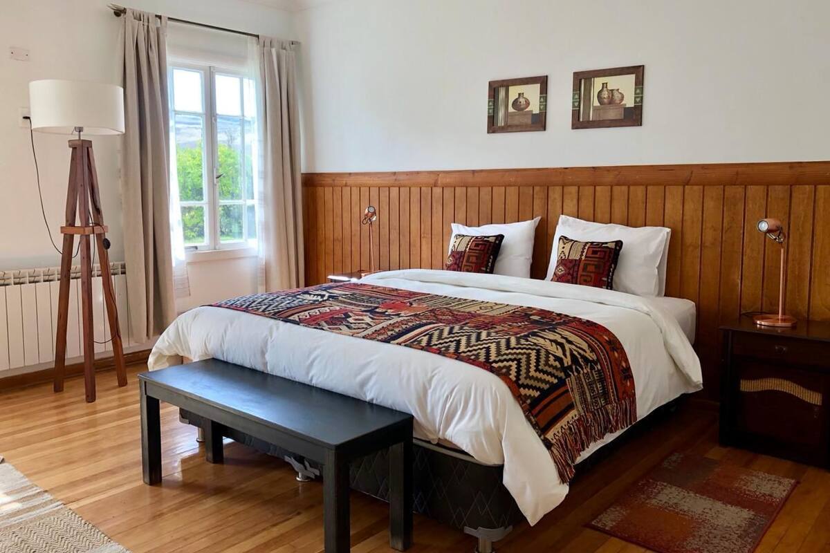 Une chambre à coucher avec une tête de lit en bois, un grand lit avec une literie à motifs, un banc, une lampe sur pied et une vue sur la verdure à l’ext érieur.