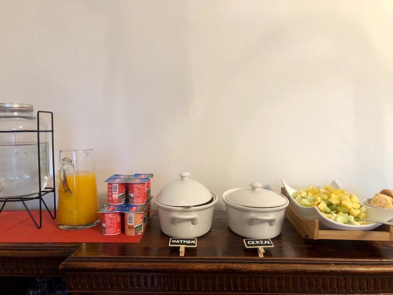 Una mesa de desayuno buffet con avena y recipientes de cereal, una jarra de jugo de naranja y una bandeja de frutas y galletas.