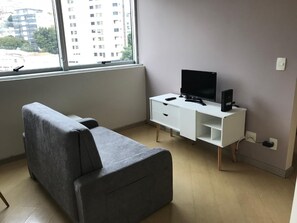 A modern room with a grey sofa, a white desk with a TV and computer, and a window with a view of buildings. - Locking Funcionarios (Belo Horizonte)