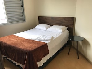 A neatly made bed with white linens and a brown throw blanket, a small round table beside it, and a window with blinds. - Locking Funcionarios (Belo Horizonte)