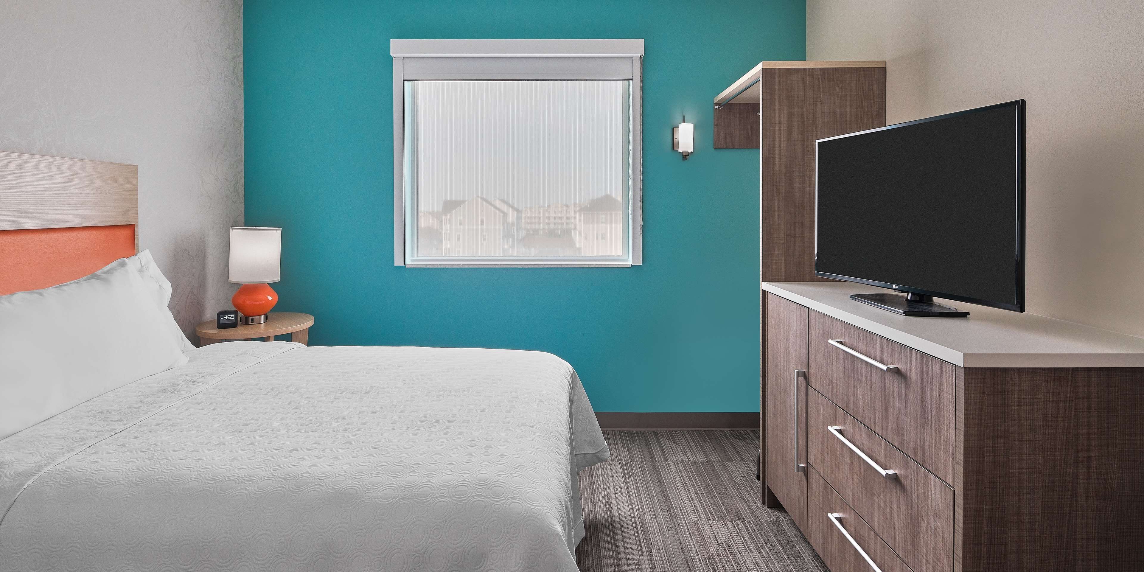 A hotel room with a bed, a TV on a cabinet, a window with a view, and a nightstand with a lamp.